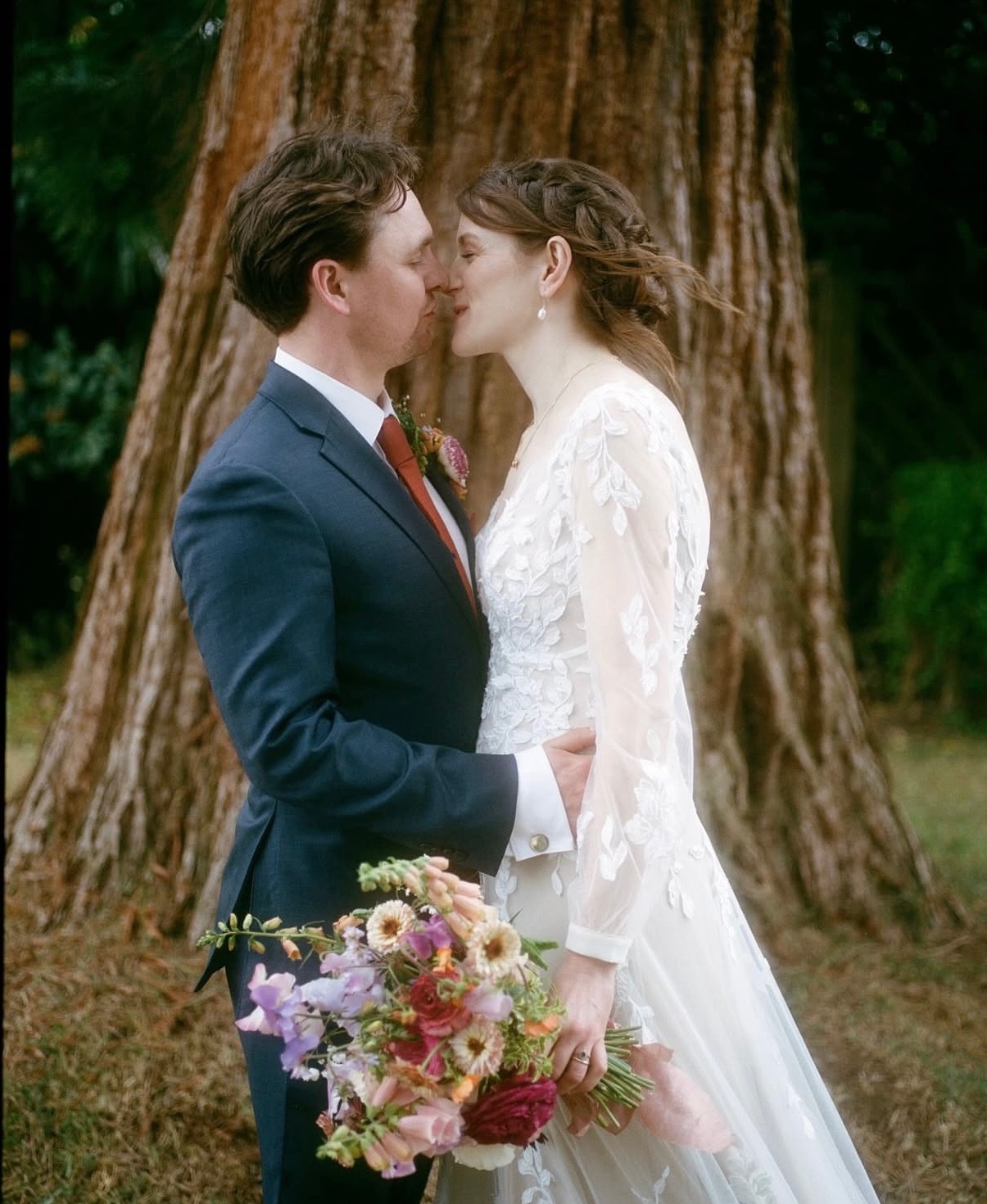 A couple sharing a kiss beneath a tree, bride holding a colourful seasonal bouquet