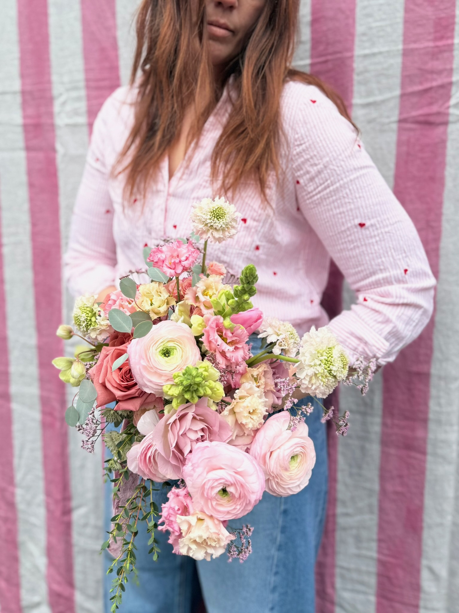 A lush pink bouquet of ranunculus, roses and eucalyptus held against a pink-striped backdrop