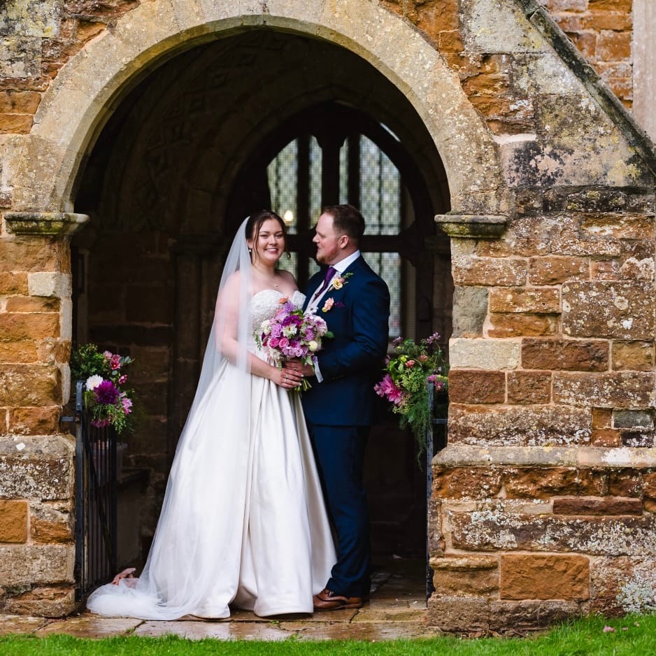 Wedding couple in a church doorway with purple and pink floral arrangements